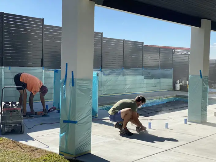 Two of the crew prepping a pool surround under an alfresco roof, glass fence and posts masked off with plastic before the overlay goes down