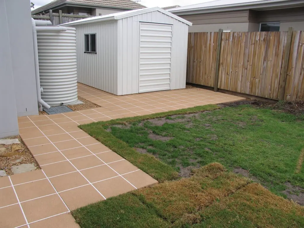 Concrete slab and path beside a garden shed and rainwater tank, finished in a tan tile grid