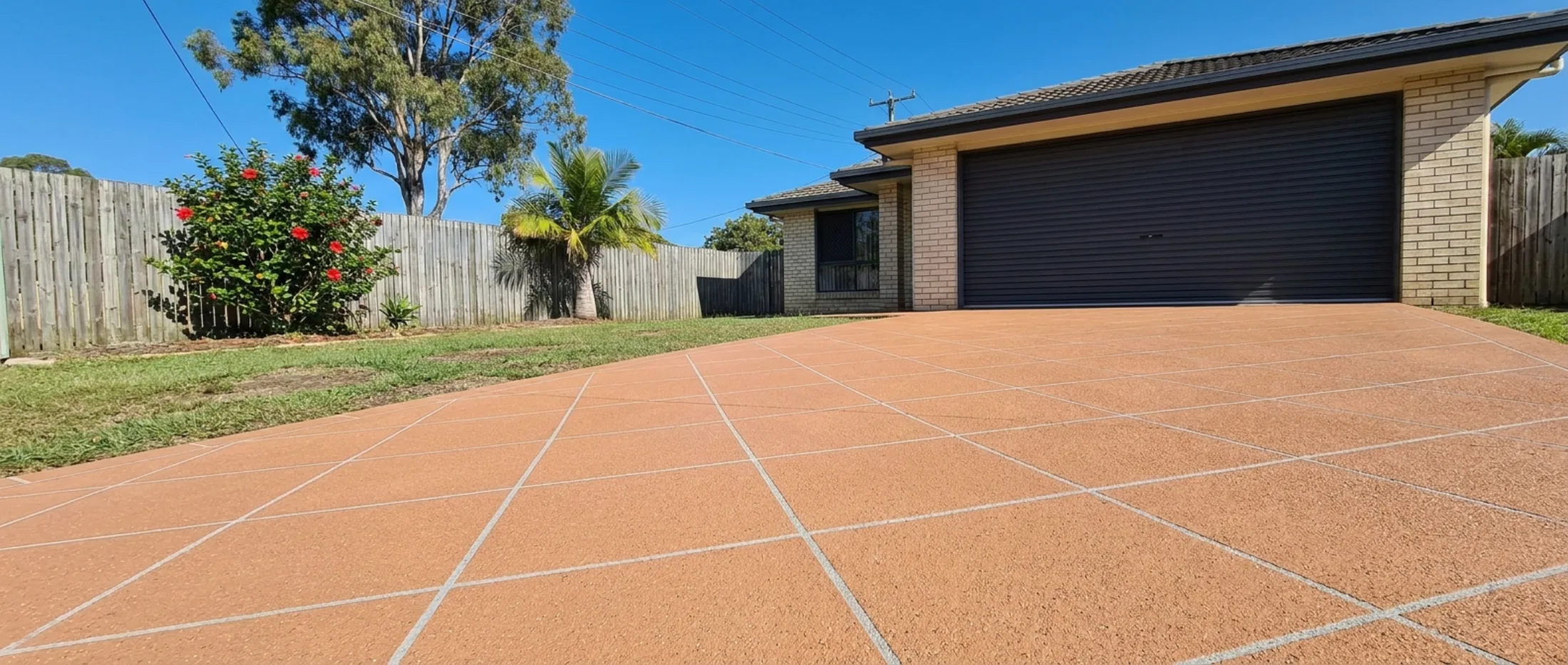 Terracotta spray-on decorative concrete driveway cut in a 45-degree diamond tile grid with white grout lines, running to the Colorbond garage of a cream brick Brisbane home under hard midday sun