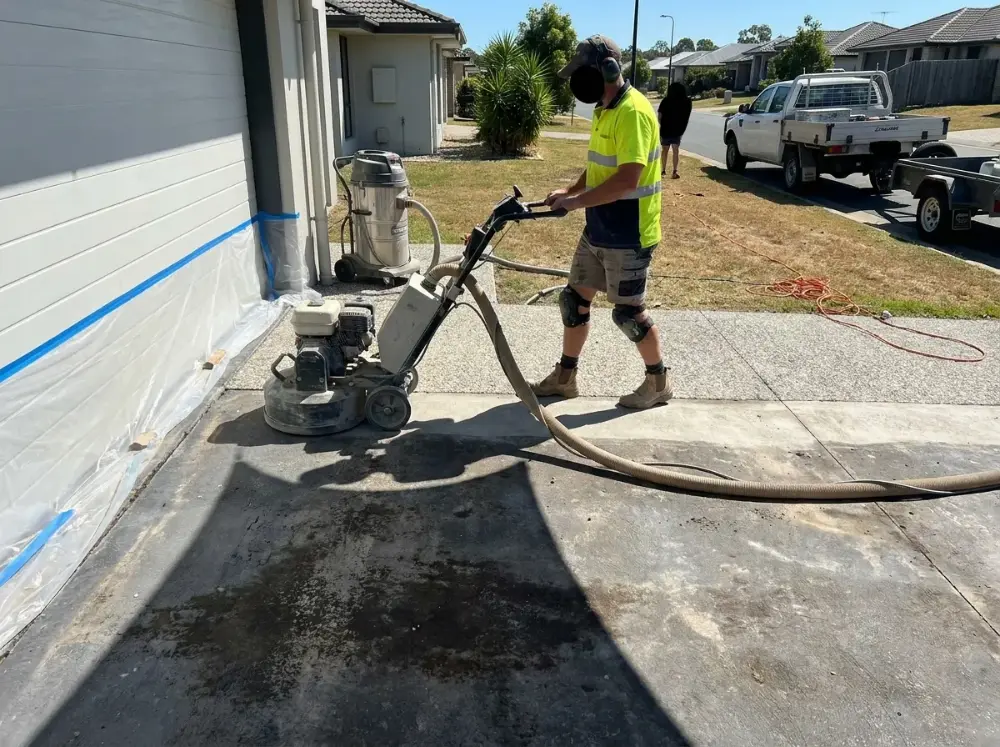 Concreter running a walk-behind diamond grinder across an old driveway slab with dust extraction